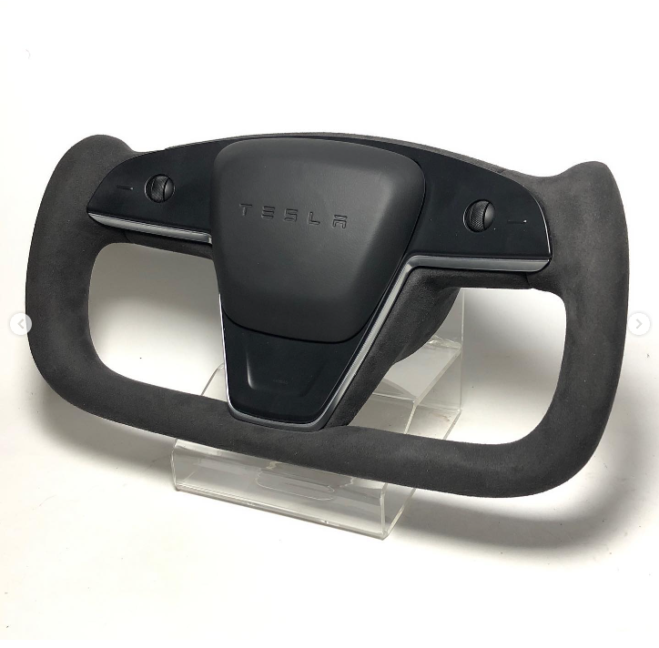A black Tesla steering wheel with a yoke design, resting on a clear stand. The center features the Tesla logo embossed, and buttons are visible on either side.