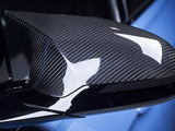 A carbon fiber car side mirror reflects light, showcasing its glossy surface and detailed texture. It is attached to a sleek, blue vehicle, emphasizing a modern, high-performance design.