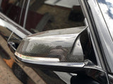 A carbon fiber car side mirror with integrated indicator light on a shiny black vehicle, parked near a brick wall reflecting the cloudy sky.