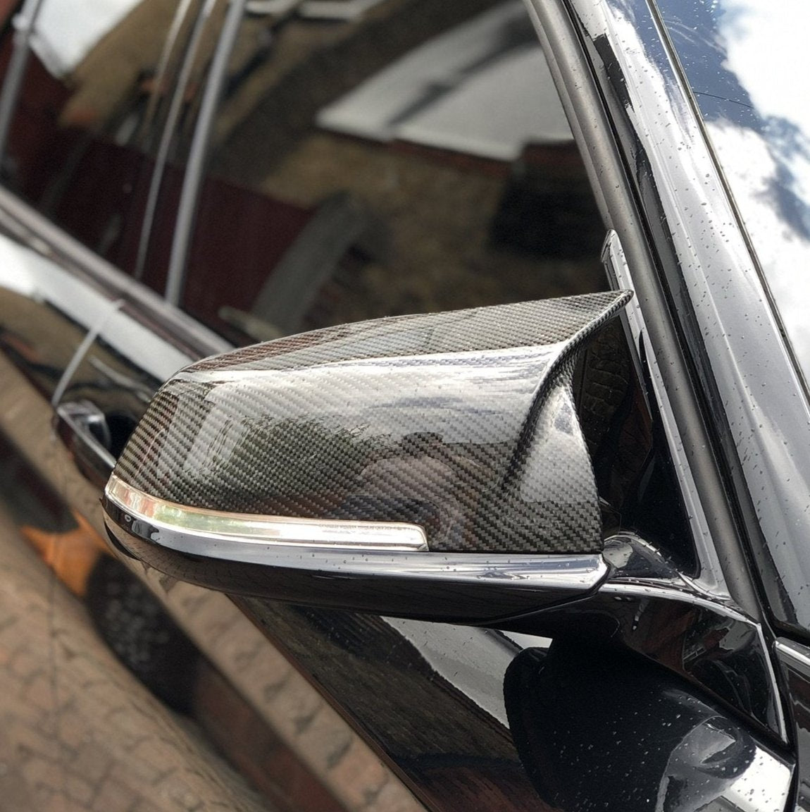 A carbon fiber car side mirror with integrated indicator light on a shiny black vehicle, parked near a brick wall reflecting the cloudy sky.