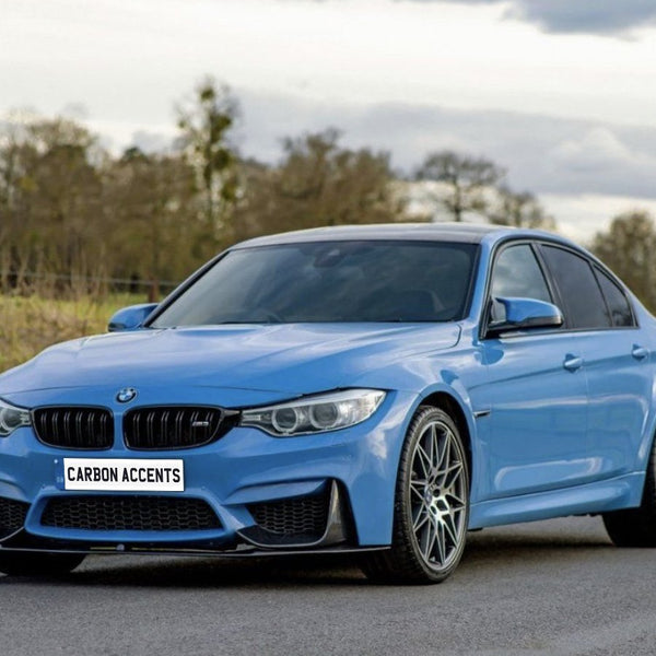 A blue sports sedan is parked on a road, with trees and clouds in the background. The license plate reads "CARBON ACCENTS."