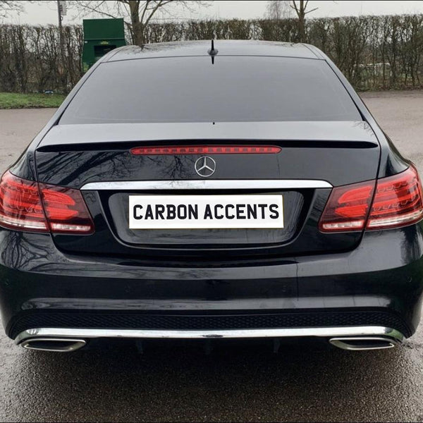 Car with black paint and a "CARBON ACCENTS" license plate remains stationary in a parking area. Background includes leafless trees and a green container.
