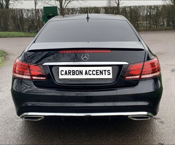 Car with black paint and a "CARBON ACCENTS" license plate remains stationary in a parking area. Background includes leafless trees and a green container.