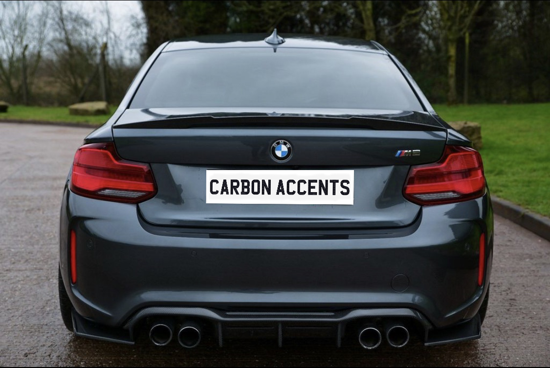 A dark gray BMW car is parked on a road. The license plate reads "CARBON ACCENTS," surrounded by a grassy area and leafless trees.