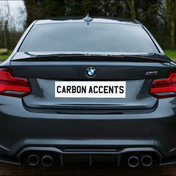 A dark gray BMW car is parked on a road. The license plate reads "CARBON ACCENTS," surrounded by a grassy area and leafless trees.