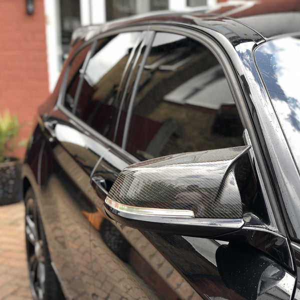 A shiny black car's side mirror reflects the sky, parked beside a red-brick building with a plant pot on a paved driveway.