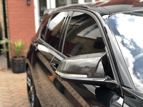 A shiny black car's side mirror reflects the sky, parked beside a red-brick building with a plant pot on a paved driveway.