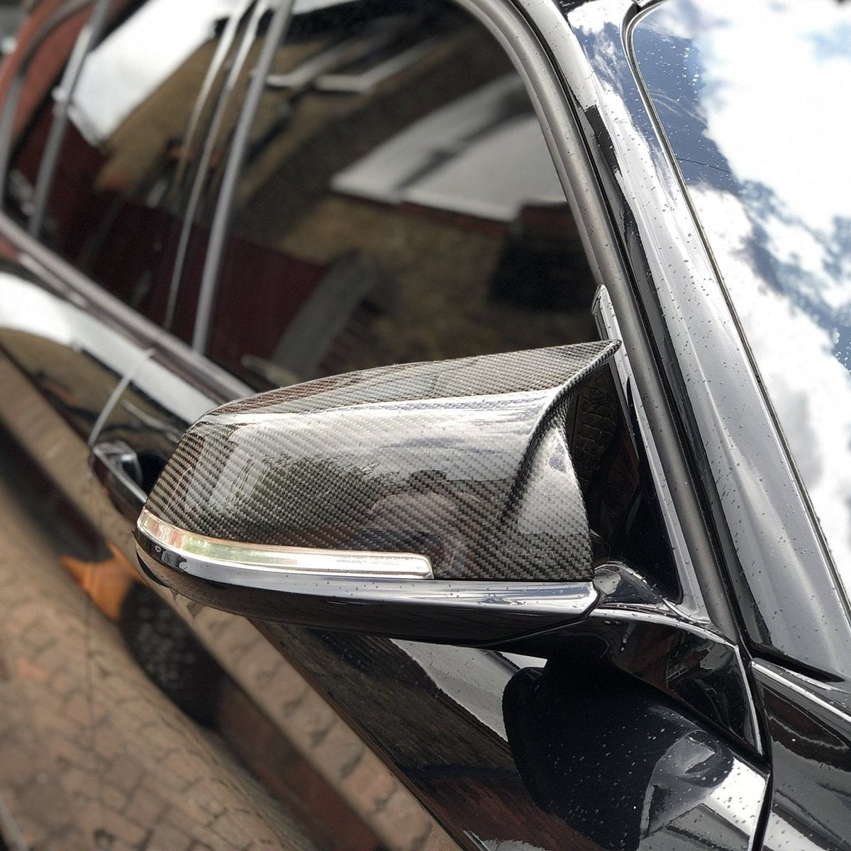 A carbon fiber side mirror is attached to a black car, reflecting buildings and sky, parked on cobblestone pavement.