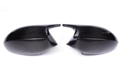 Two black carbon fiber car mirror covers face each other, resting on a plain white background, highlighting their glossy texture and sleek design.