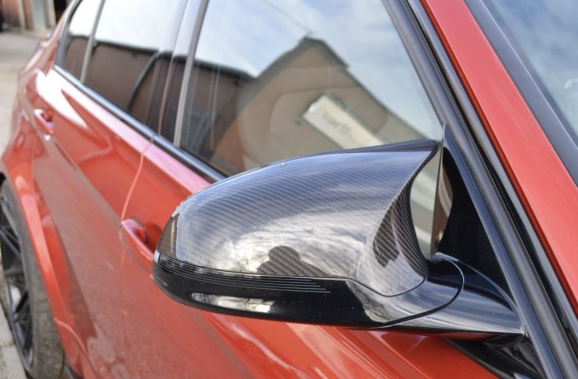 Carbon fiber side mirror on a red car reflects the sky and nearby buildings, parked outside an industrial area with brick structures.