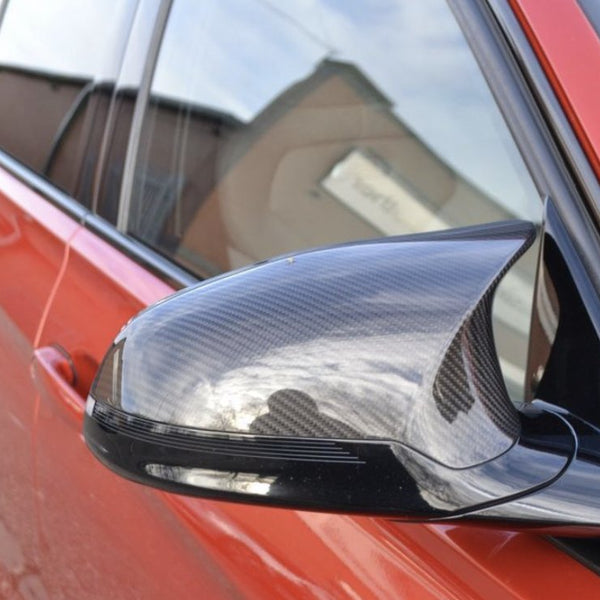 Carbon fiber side mirror on a red car reflects the sky and nearby buildings, parked outside an industrial area with brick structures.