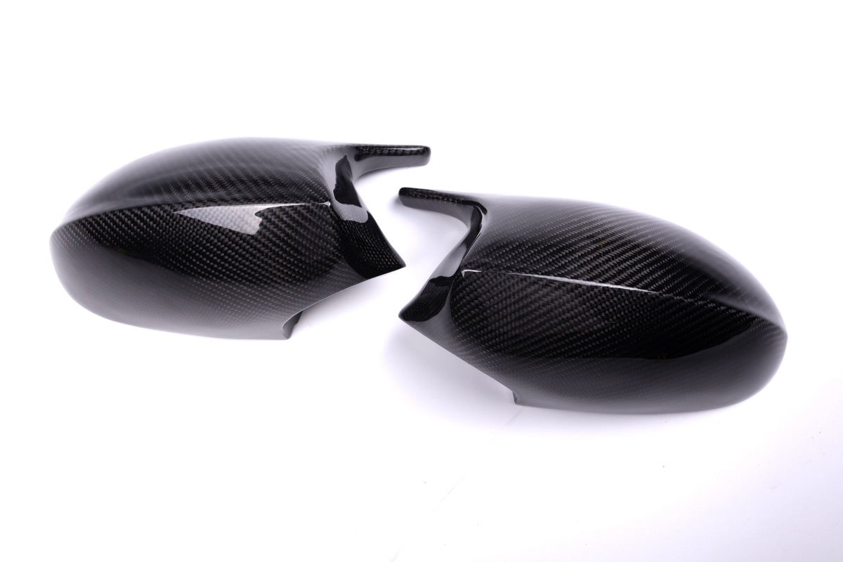 Two sleek carbon fiber side mirror covers, positioned facing each other, rest against a plain white background. Their dark, glossy surface highlights the distinctive carbon fiber texture.