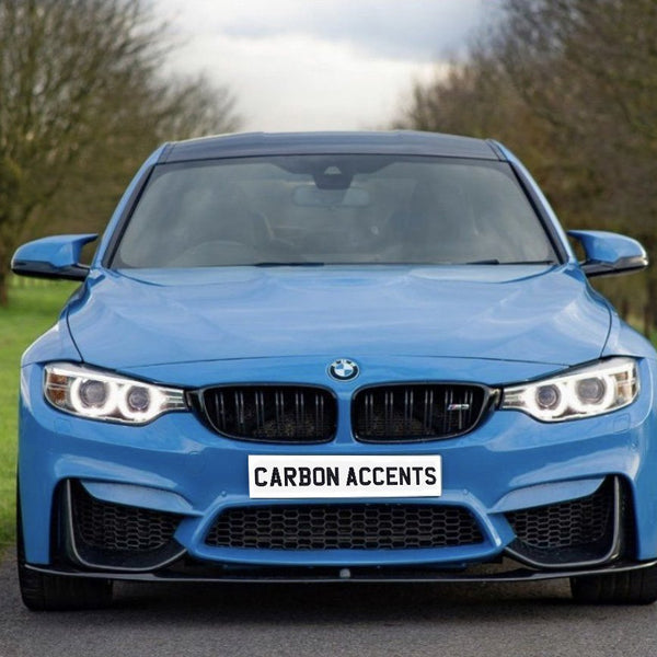 A blue car is parked on a rural road, with trees lining the background. The front license plate reads "CARBON ACCENTS." The car's headlights are illuminated.