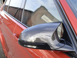 A car's side mirror, featuring a carbon fiber texture, reflects a cloudy sky. The vibrant red vehicle is parked in an outdoor setting, with a building visible in the background.