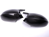 Two carbon fiber mirror caps rest on a white surface, featuring a glossy finish and textured pattern, suggesting use in automotive applications.