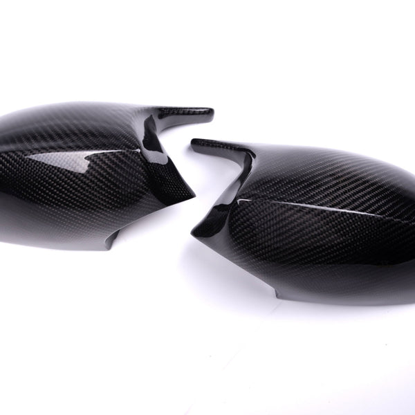 Two carbon fiber mirror caps rest on a white surface, featuring a glossy finish and textured pattern, suggesting use in automotive applications.