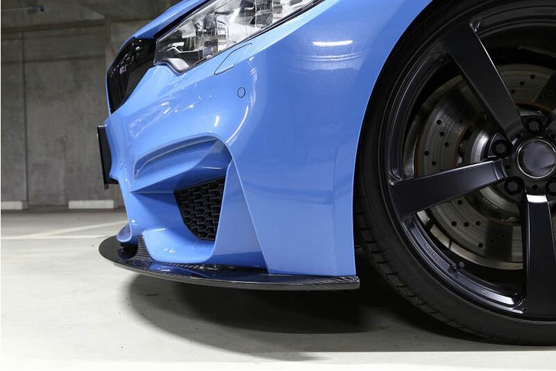 A blue sports car, parked in a concrete garage, displays its front bumper, headlight, and a large, glossy black alloy wheel with visible brake disc.