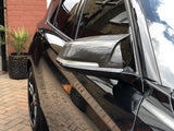 A black car with a carbon fiber side mirror is parked on a brick driveway, reflecting a building. Nearby are potted plants and a brick wall with a white-framed door.