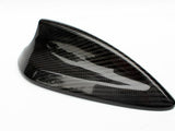 A sleek, black carbon-fiber car antenna rests on a white surface, showcasing a streamlined, shark-fin design that reflects light, highlighting its glossy texture and modern aesthetics.
