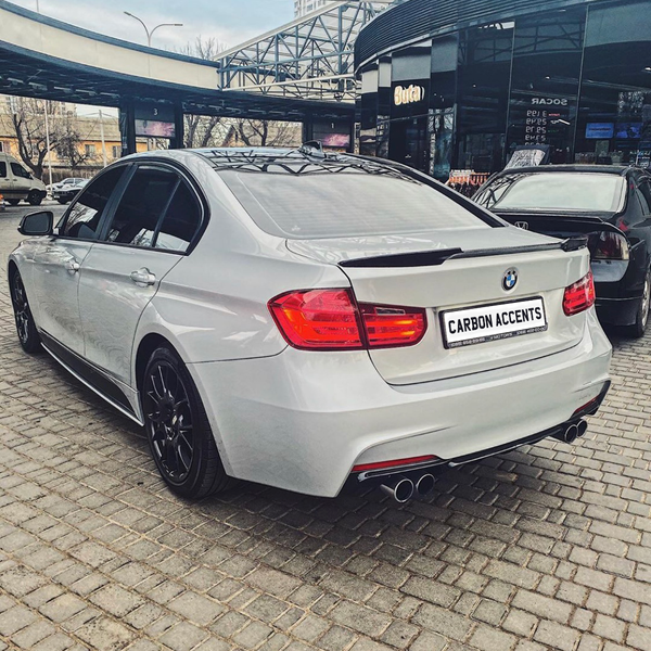 A silver BMW sedan is parked on a cobblestone area near a storefront labeled "Buta" with a "CARBON ACCENTS" license plate, surrounded by other vehicles and structures.