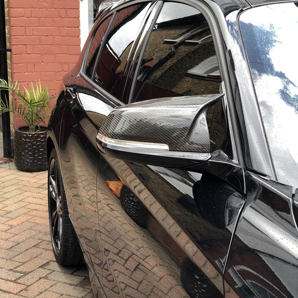 A black car with tinted windows and carbon fiber side mirror is parked on a brick driveway. Its reflective surface displays a brick wall and glass doors in an urban setting.