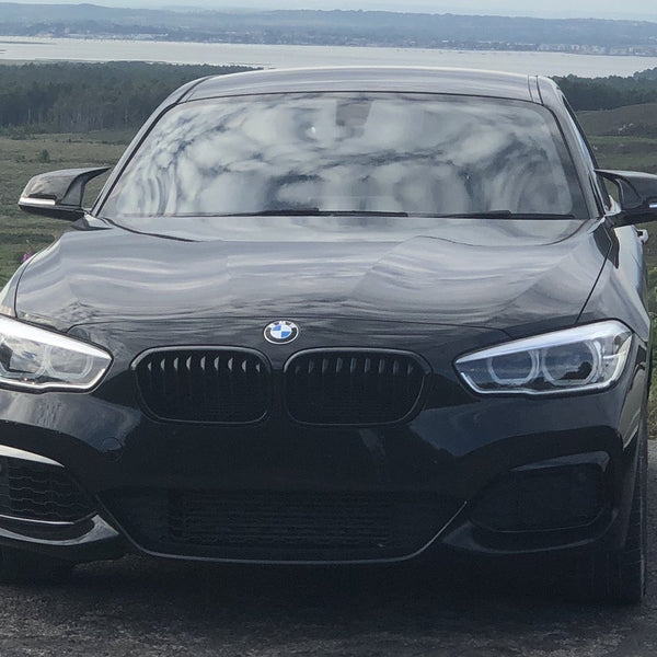 A black BMW car is parked on a rural road with greenery and hills stretching to a distant body of water, under a cloudy sky.