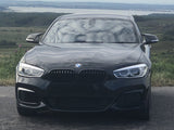 A black BMW car is parked on a road, facing forward, with rolling hills and a body of water in the distant background.