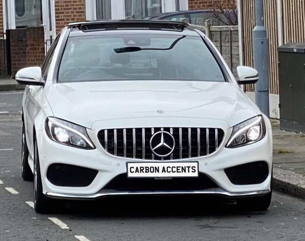 Car parked on a residential street, with LED headlights on. It's a white Mercedes-Benz with distinctive grille and license plate reading "CARBON ACCENTS." Bricks and lampposts are visible nearby.