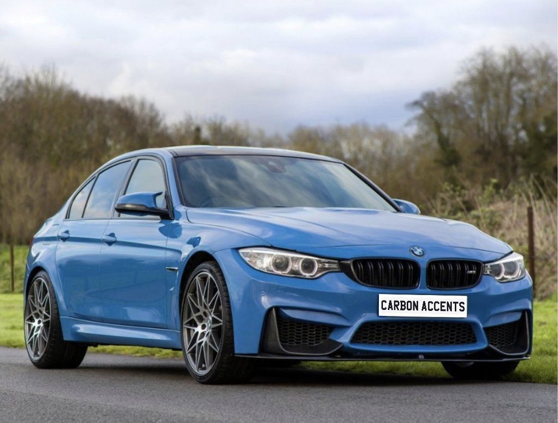 A blue sports car is parked on a road, displaying "CARBON ACCENTS" on the license plate. The background features a grassy area and leafless trees under a cloudy sky.