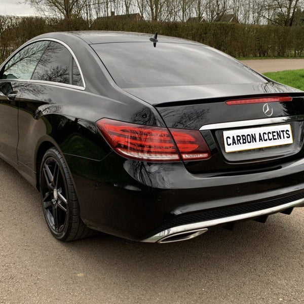 A black luxury car is parked on a road, with "CARBON ACCENTS" on the license plate. The car is set against a backdrop of grass and trees.