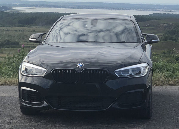 A black BMW car is parked on a road, facing forward, with a hilly landscape and a large body of water in the background.
