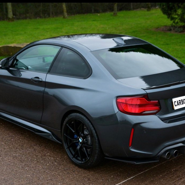 A dark gray sportscar with tinted windows is parked on a wet road. The license plate reads "CARBON ACCENTS," and lush green grass borders the scene.