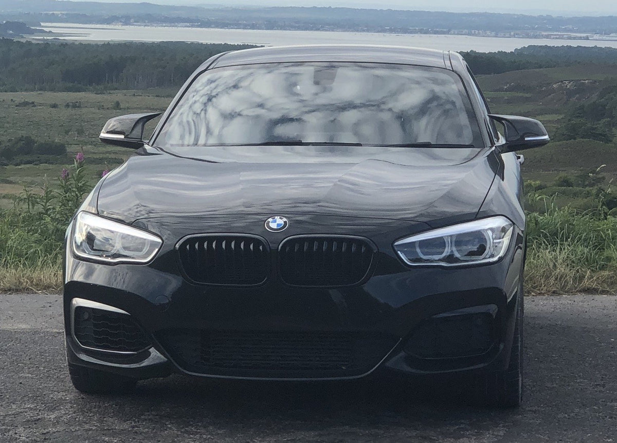 A black BMW car is parked on a road, facing forward, with a scenic backdrop of fields and a distant body of water under a cloudy sky.