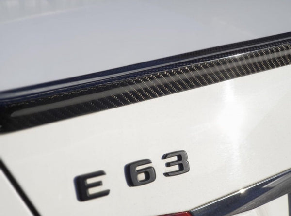 The image shows the rear emblem "E 63" on a white car, featuring a carbon fiber detail on the trunk lid in bright sunlight.