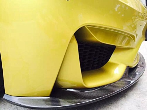 A yellow car's front bumper features a black carbon fiber splitter, providing aerodynamic enhancement. The context is a close-up view, emphasizing design details on the ground level.