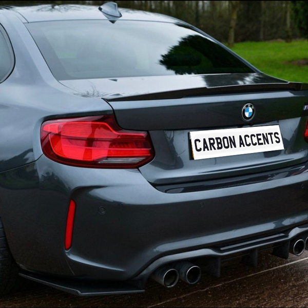 A dark gray BMW sports car is parked on a wet road, featuring a dual exhaust and distinctive rear lights, with a license plate reading "CARBON ACCENTS" in a green, grassy environment.