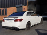 A white Mercedes-Benz E63 AMG is parked on a paved area outside an industrial building with orange brick walls. The text 