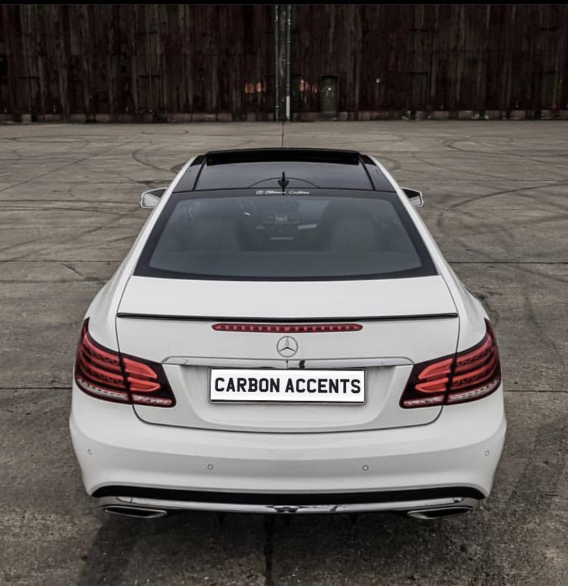 A white Mercedes-Benz car is parked on a concrete surface. The license plate reads "CARBON ACCENTS" against a wooden backdrop.