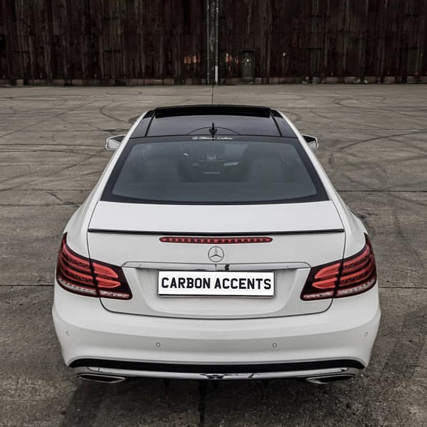 A white Mercedes-Benz car is parked on a concrete surface. The license plate reads "CARBON ACCENTS" against a wooden backdrop.