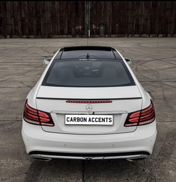 A white Mercedes-Benz car is parked on a concrete surface. The license plate reads "CARBON ACCENTS" against a wooden backdrop.