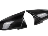 Two sleek, carbon fiber side mirror covers are angled toward each other, showcasing a reflective surface and intricate weave pattern against a plain white background.