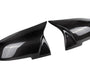 Two black carbon fiber car mirror covers are displayed, facing each other. They have a glossy finish and reflect light, emphasizing the smooth, aerodynamic design against a plain white background.