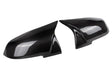 Two black carbon fiber car mirror covers are displayed, facing each other. They have a glossy finish and reflect light, emphasizing the smooth, aerodynamic design against a plain white background.