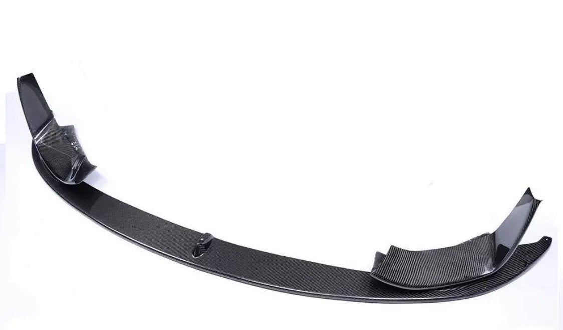 Carbon fiber car bumper lip sits on a plain white surface, showcasing its curved design and textured finish, with raised ends for aerodynamic improvement.