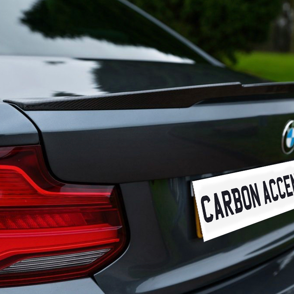 A car's rear shows a sleek spoiler and a visible logo, with text reading "CARBON ACCENTS" on the license plate. It's parked on a driveway beside green grass.