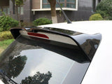 A car rear end with a spoiler and antenna is parked outdoors near a building, surrounded by greenery and trees. Reflections are visible on the rear window.