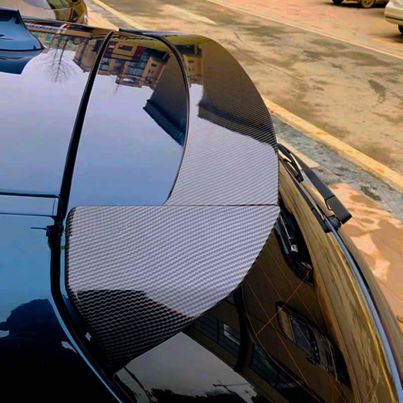 A carbon fiber car spoiler is mounted on a sleek black vehicle, reflecting nearby buildings and parked along a street with another car in the background.