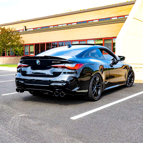 A sleek black BMW M440i is parked diagonally in a nearly empty parking lot, with a brick building featuring red-framed windows in the background.