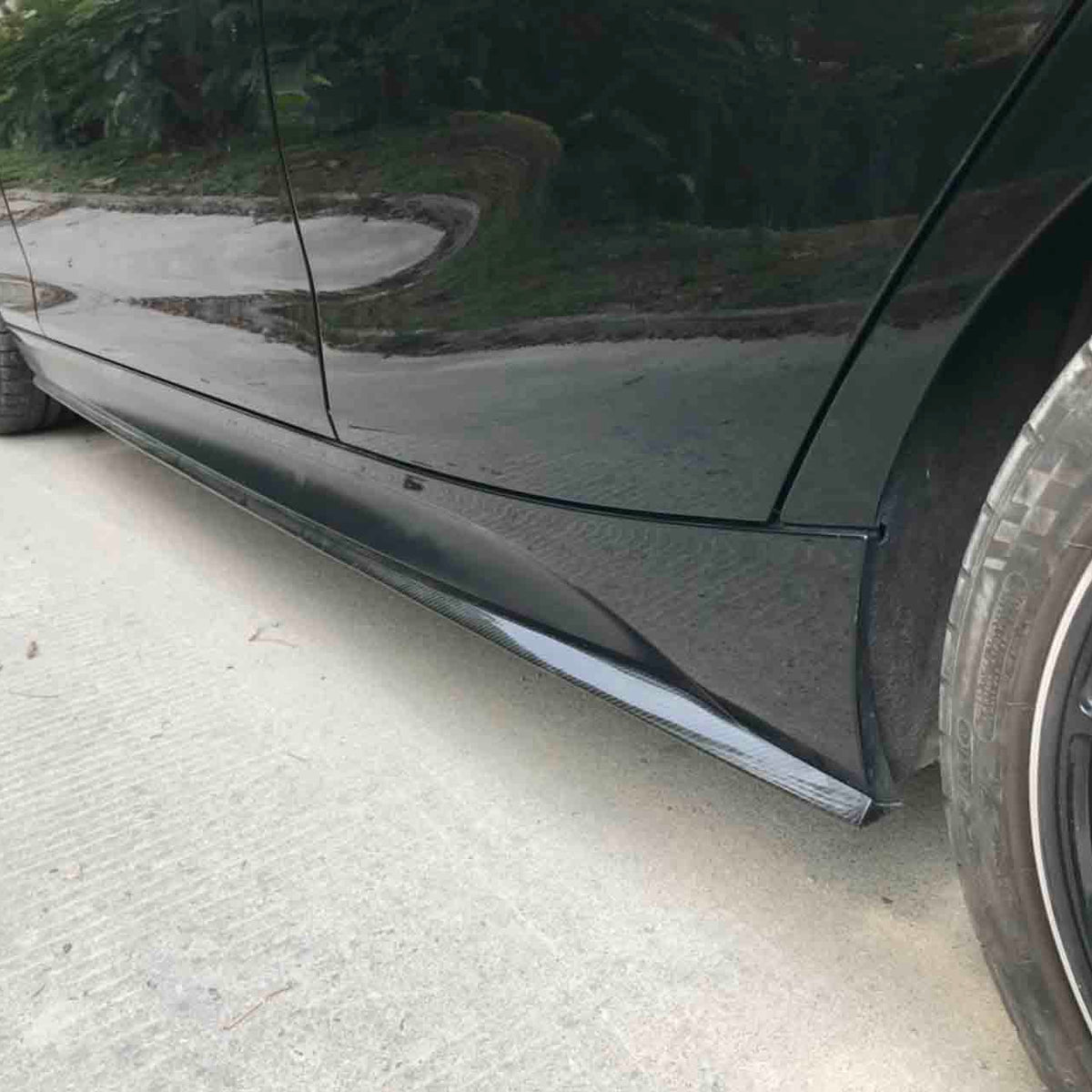 Car side skirts run along the bottom edge, enhancing aerodynamics, with black wheels on smooth pavement. Greenery is visible in the background.