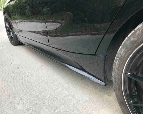 Car side skirts run along the bottom edge, enhancing aerodynamics, with black wheels on smooth pavement. Greenery is visible in the background.